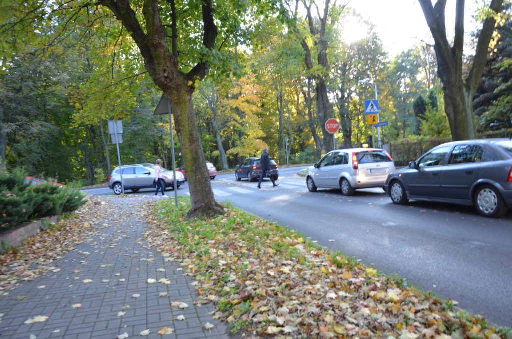 ulica, przejscie dla pieszych , dwie osoby przechodzą przez przejście, widaoczne sa trzy samochody na jezdni,