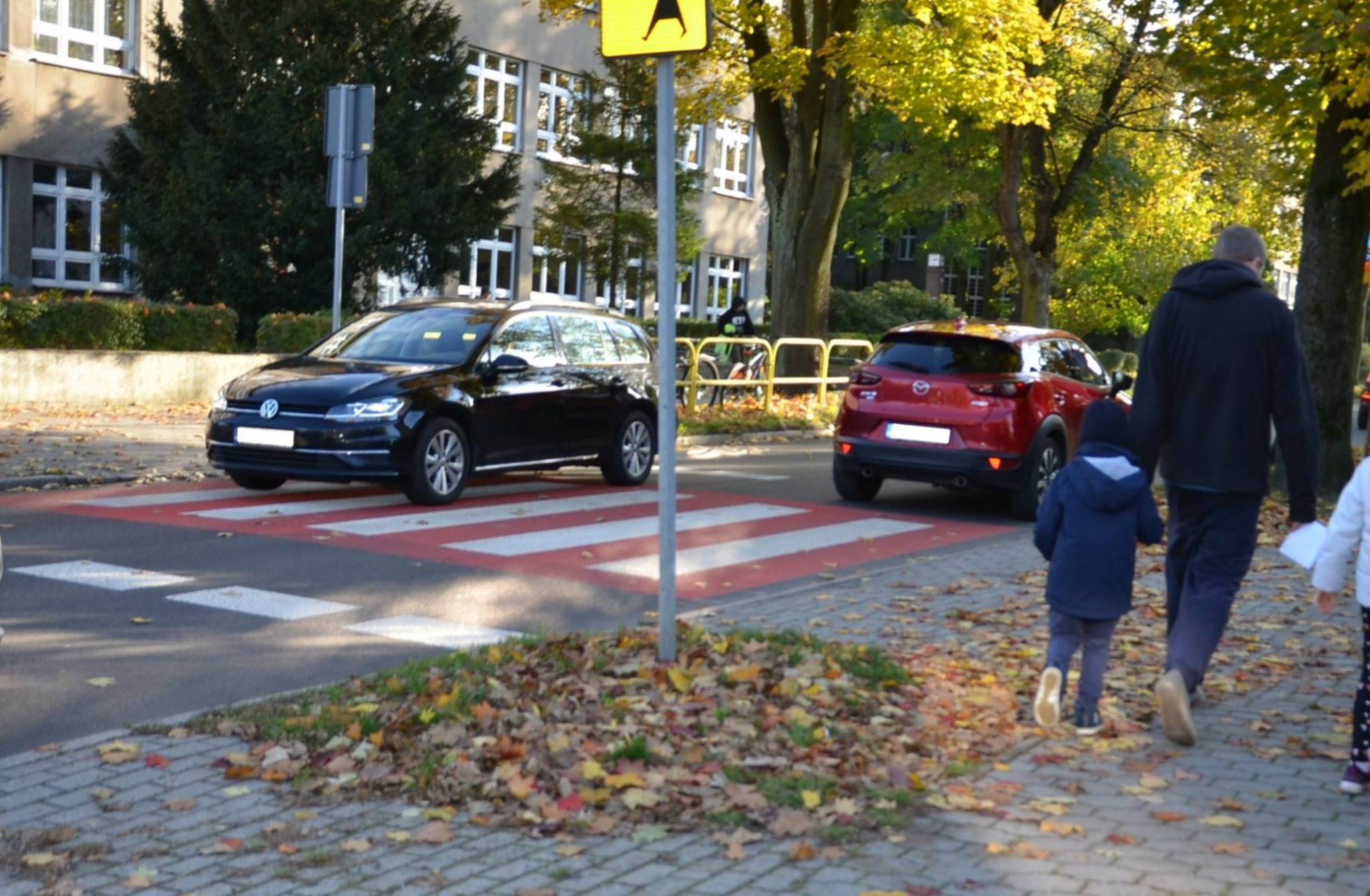 dwa samochody na jezdni, obok idą dzieci chodnikiem, po lewej stronie przejście dla pieszych,