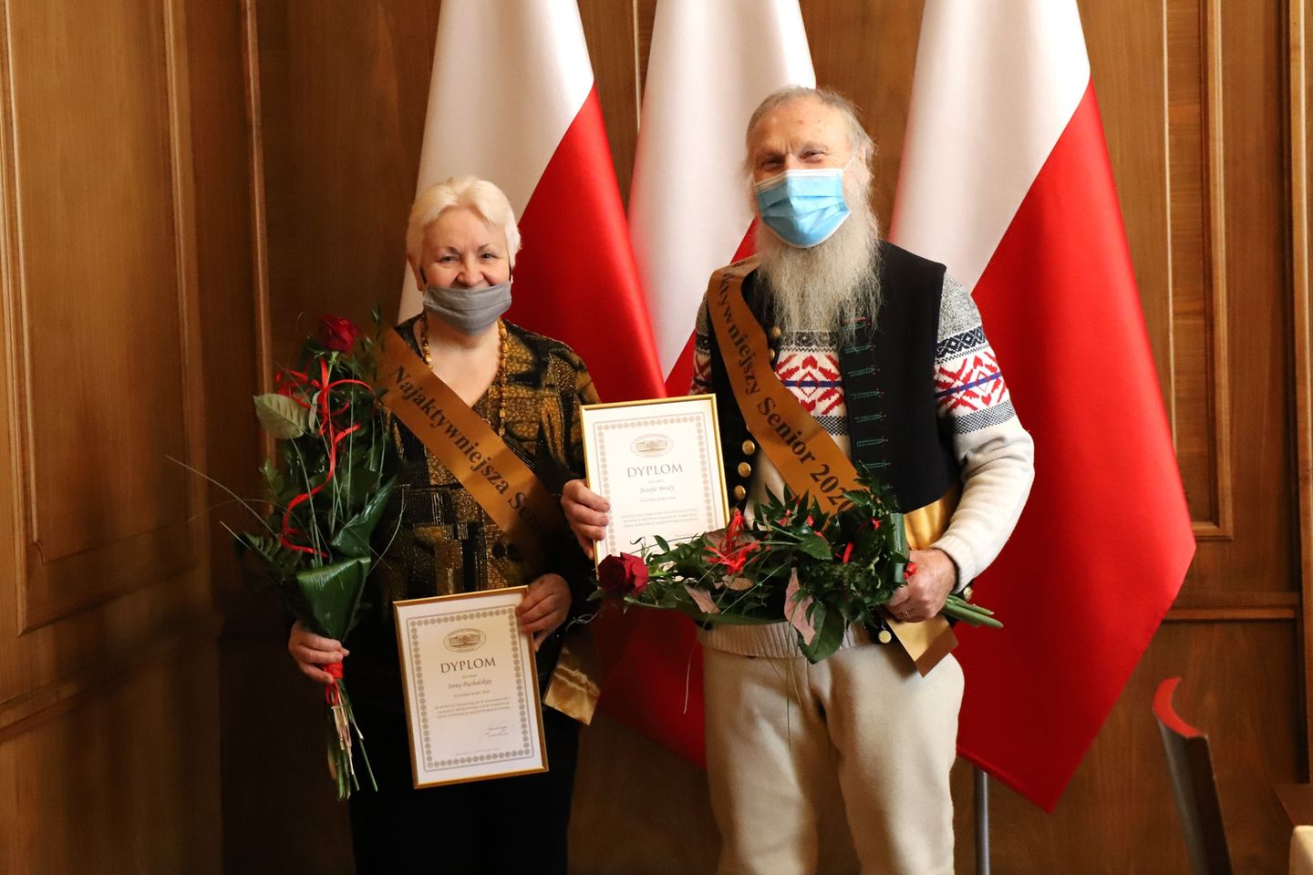 konieta i mężczyzna stoja z rękach trzymają dyplomy i kwiaty, w tle falgi państwowe Polski
