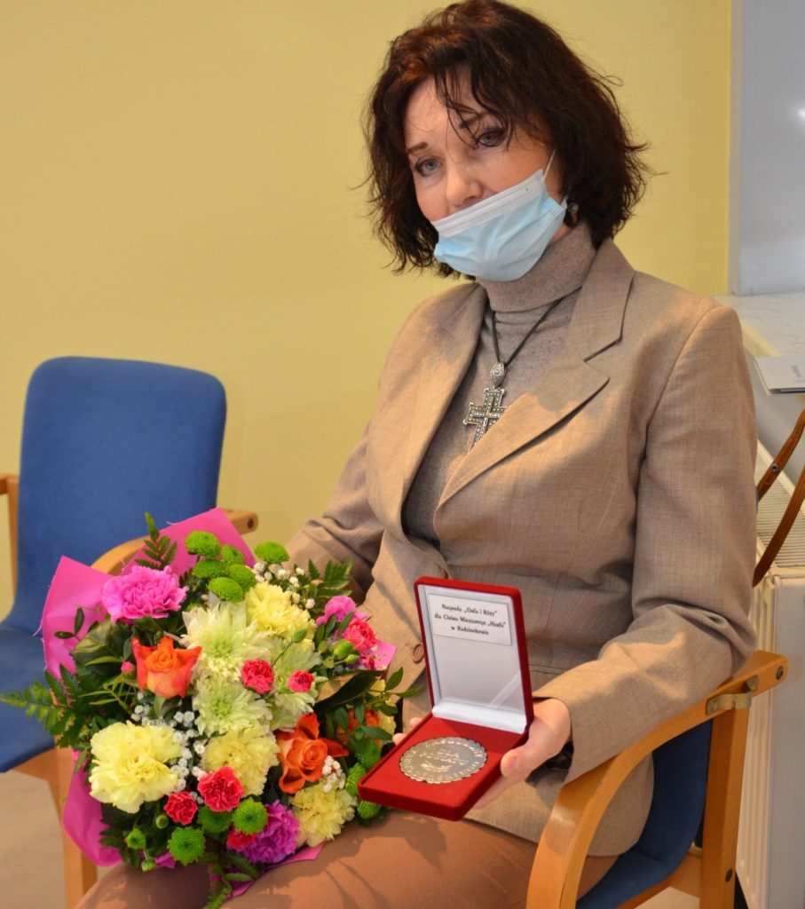 kobieta siedzi na krześle, w trzyma bukiet kwiatów oraz medal, odznaczenie Orła i Róży