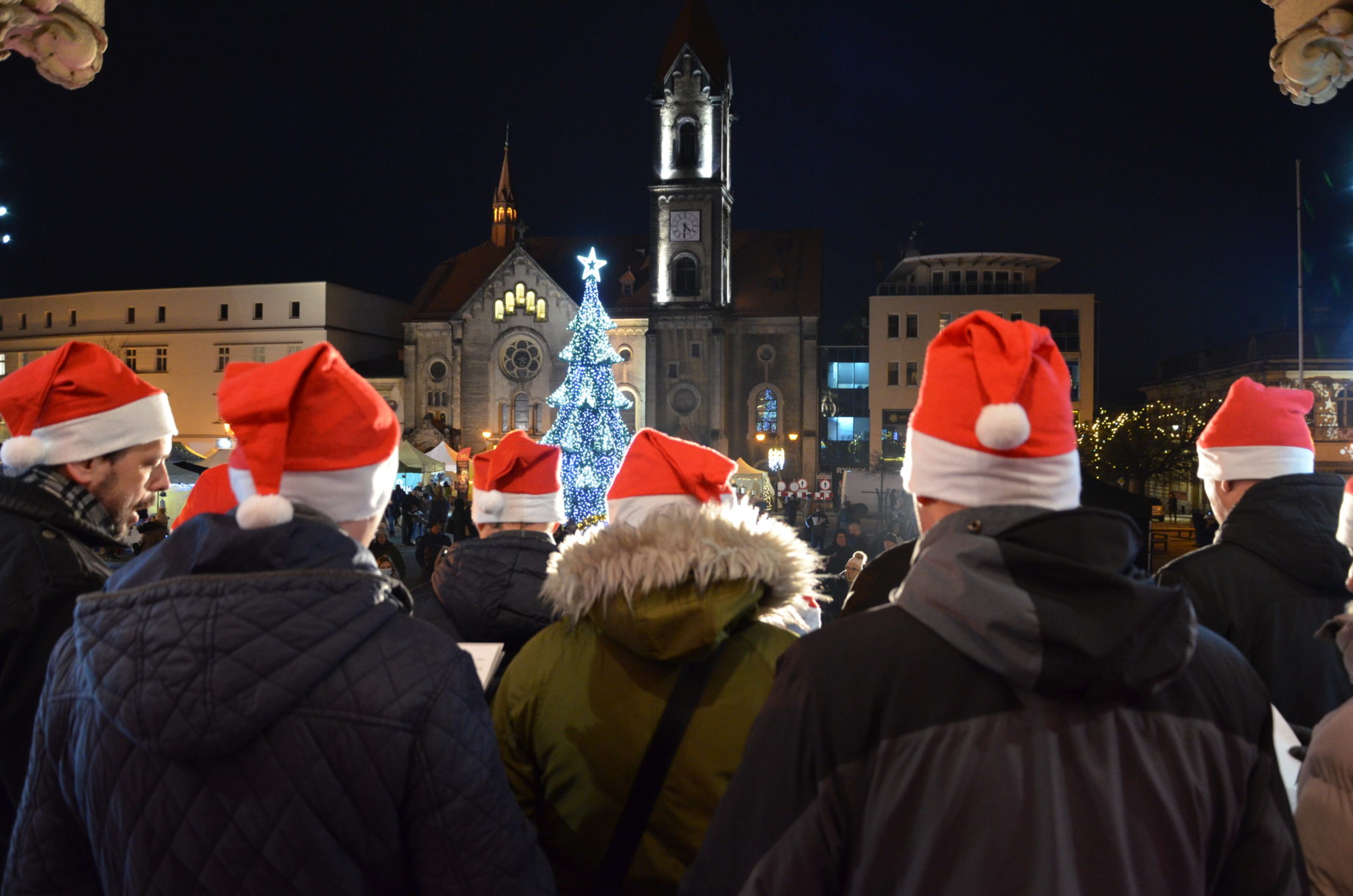 osoby w czapkach Mikołaja stoja tyłem , na drugim planie choinka