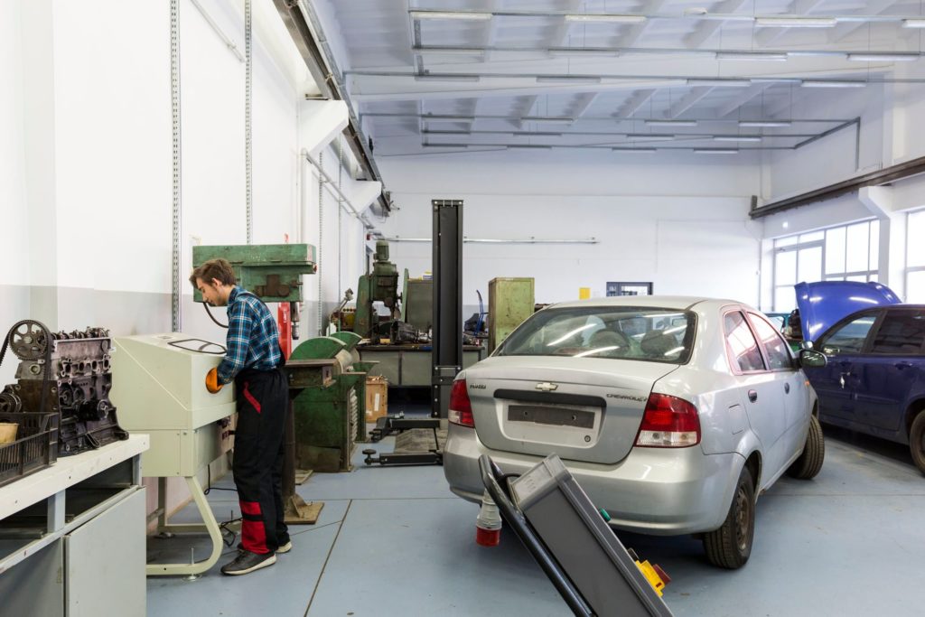 młody mechanik stopi obok stanowiska roboczego na warsztacie samochodwym, obok srebrny samochód osobowy
