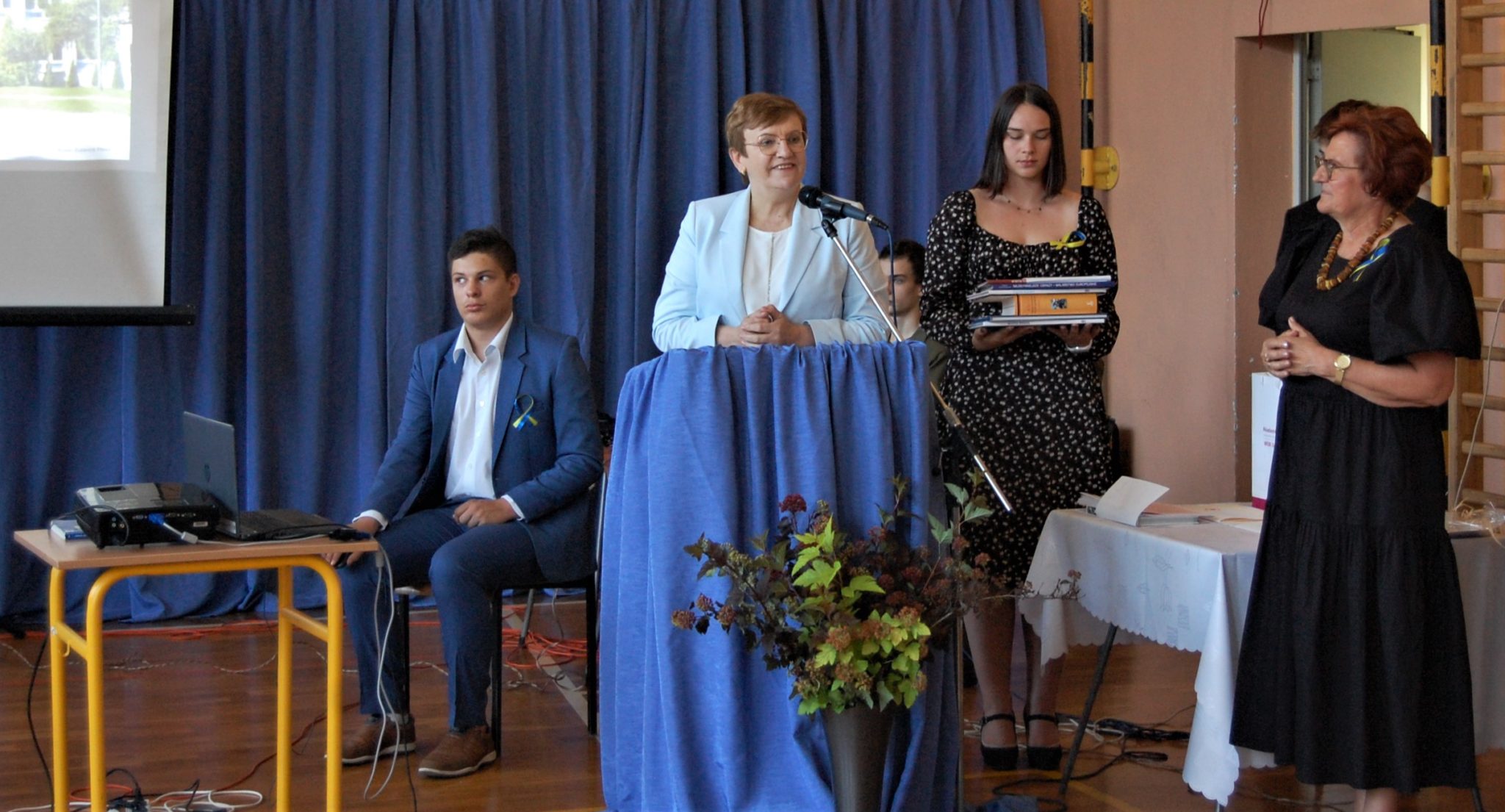 kobieta stojąca przy mównicy, obok niej stoją dwie kobiety i siedzi na stołku mężczyzna
