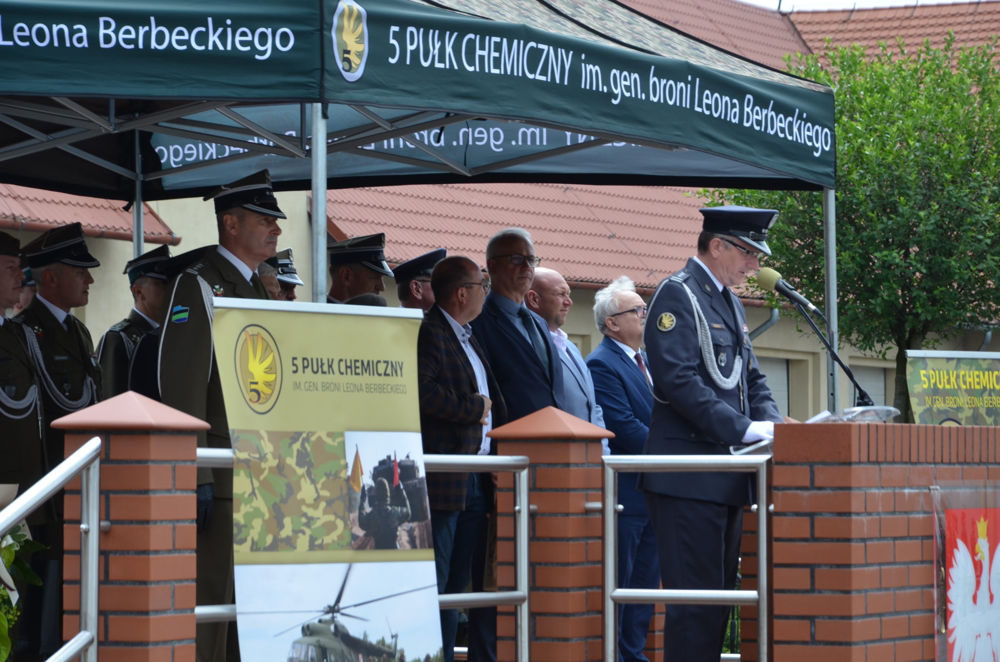 grupa ludzi zgromadzona pod zadaszeniem, z prawej strony mężczyzna stojący przy statywie z mikrofonem