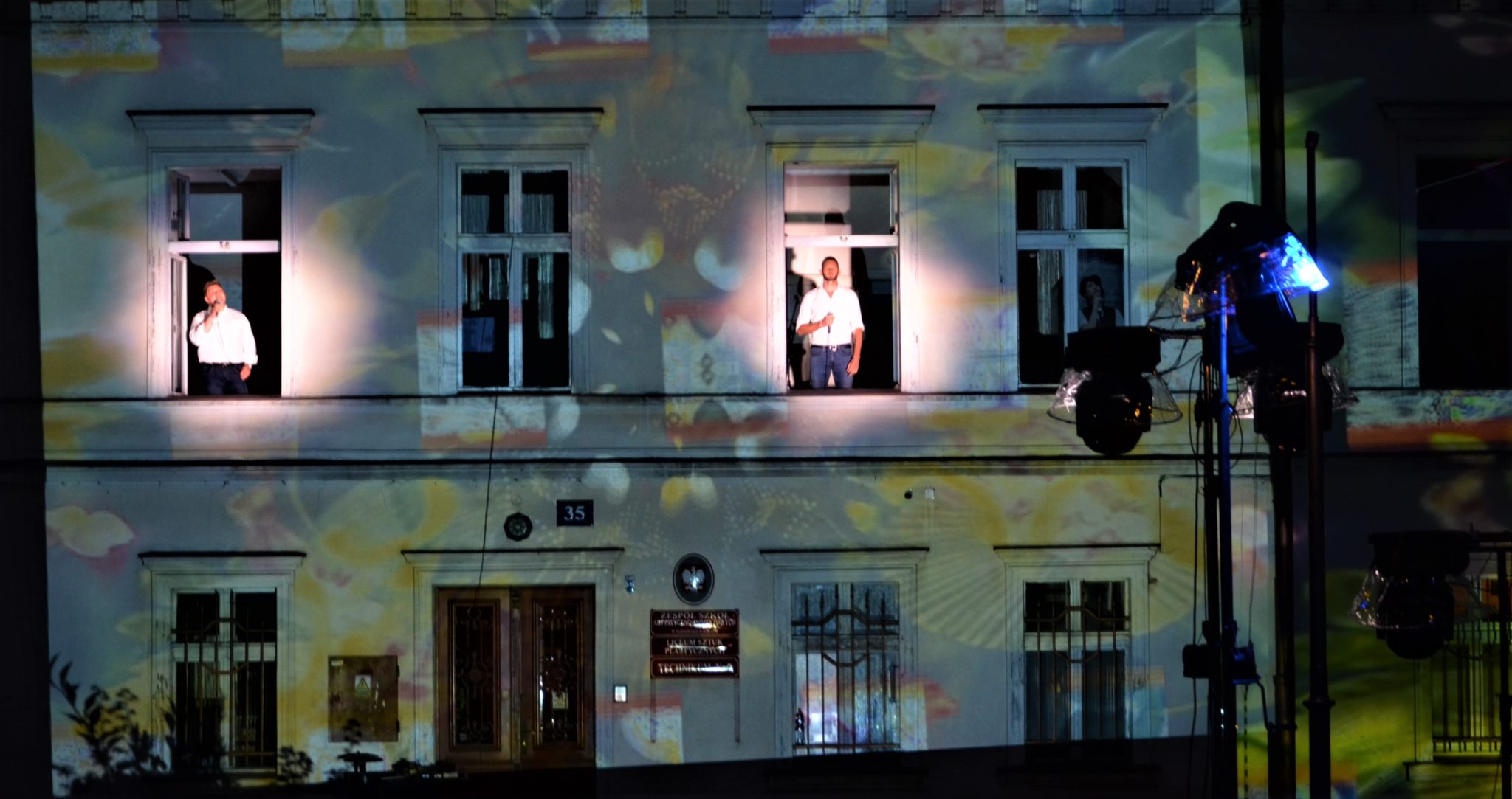 ściana budynku z iluminacją świetlną, w dwóch oknach znajdują się stojący mężczyźni