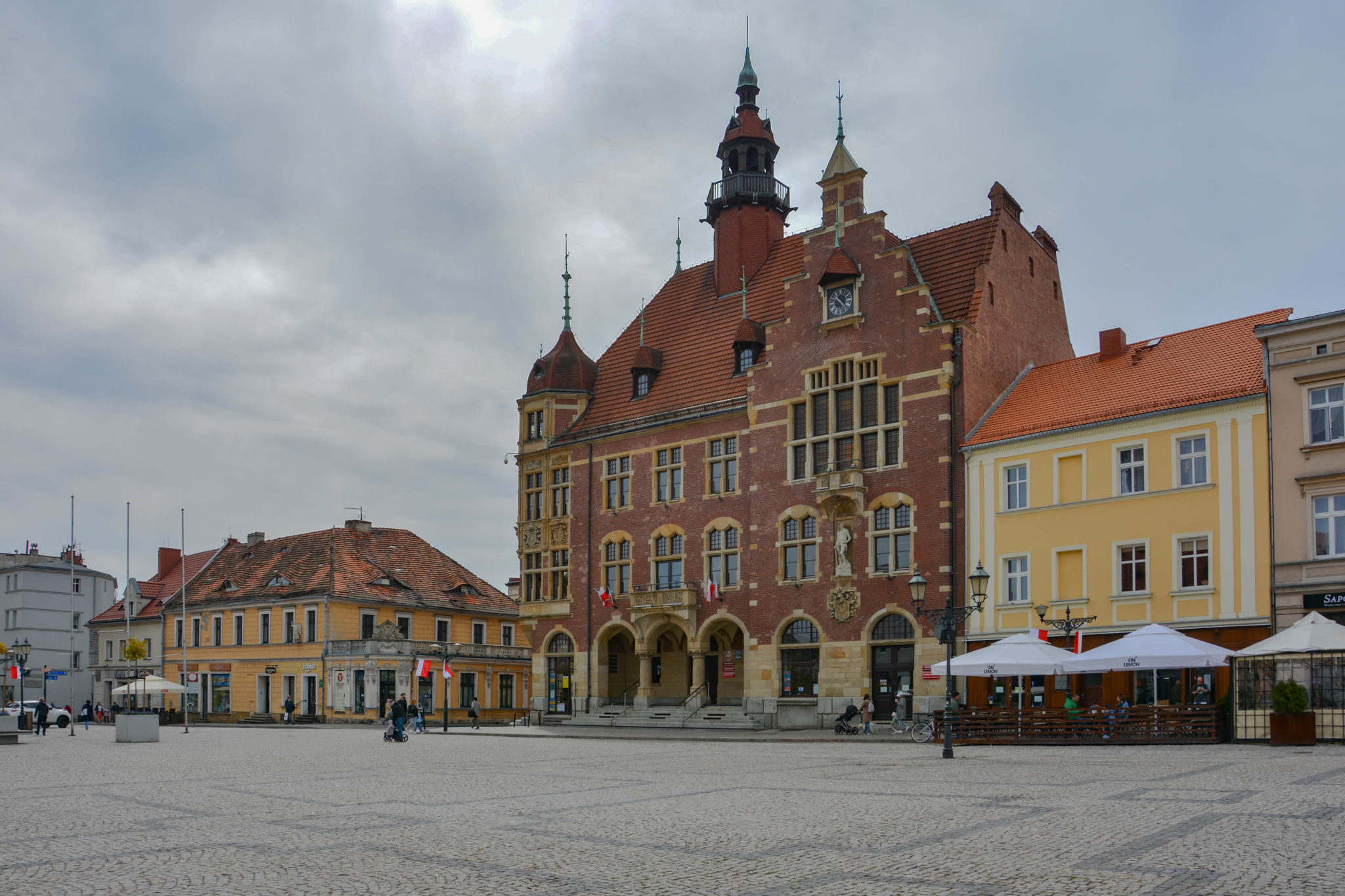 Na pierwszym planie plac z nawierzchni brukowej, w tle zabudowa o różnej wysokości, dominujący budynek z czerwonej cegły