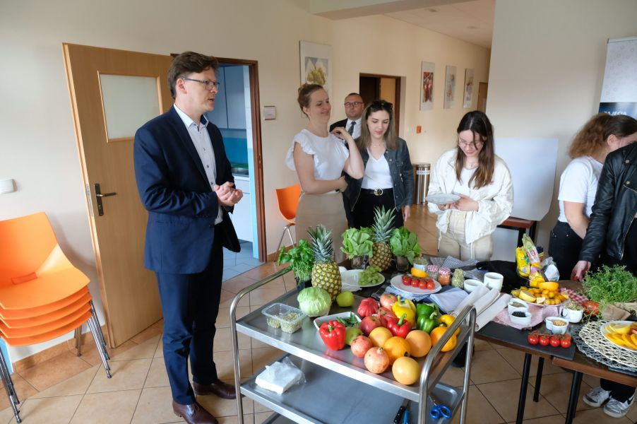 grupa osób stojących przy stoliku, na którym znajdują się warzywa, owoce i przybory kuchenne