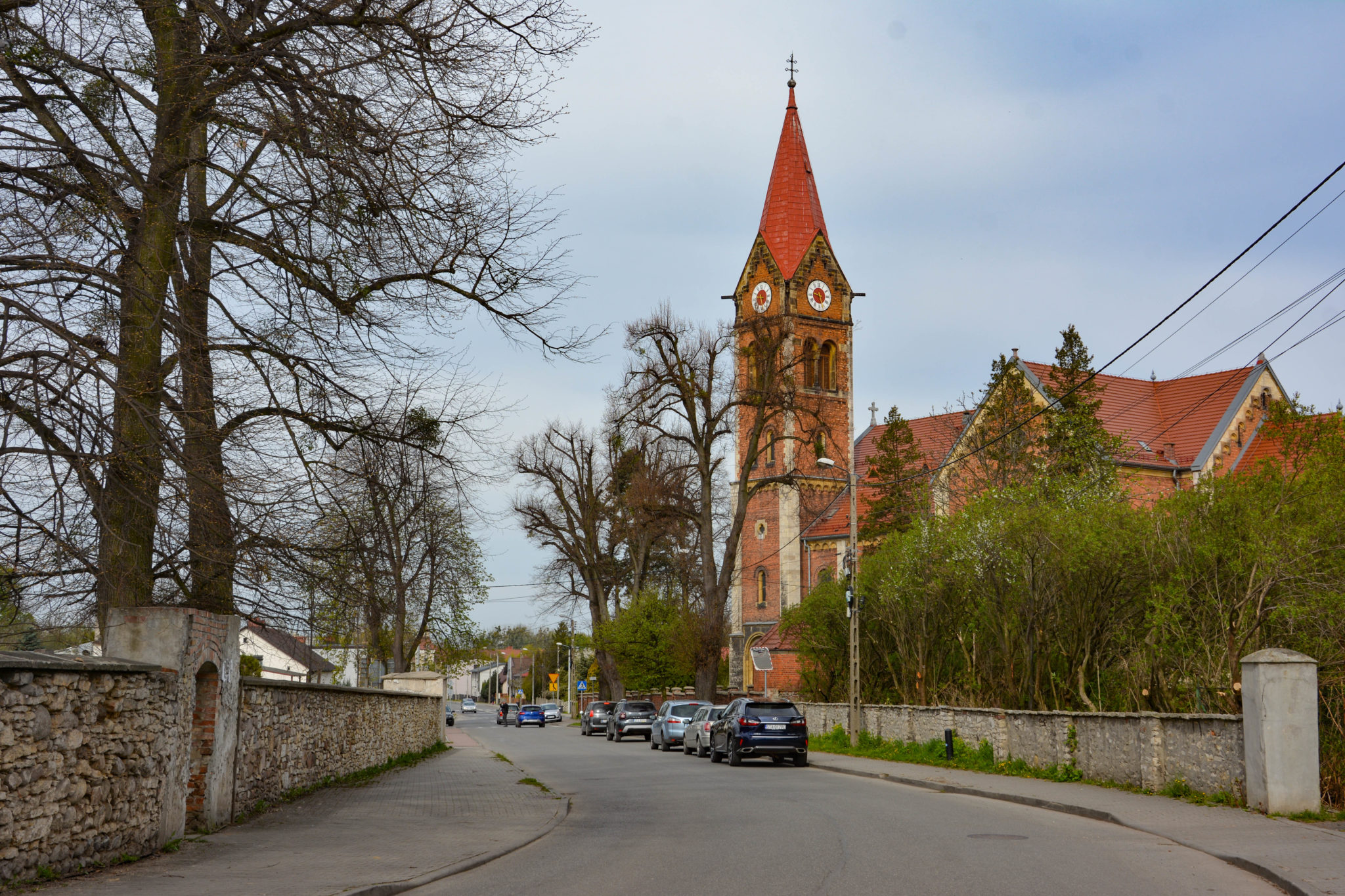 widok ulicy, po prawej stronie zaparkowane samochody osobowe, w tle wieża kościołu