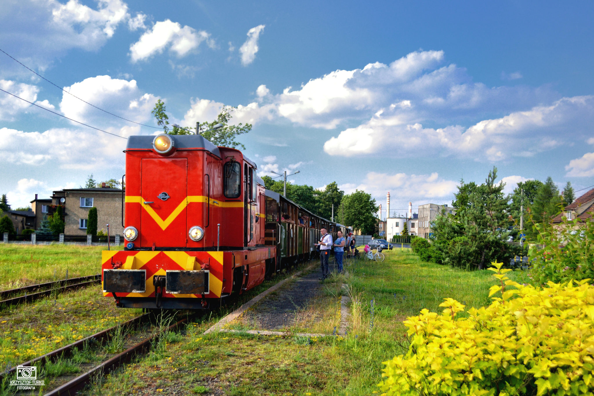 czerwony pociąg znajdujący się na torach, po prawej stronie osoby stojące na terenie zieleni