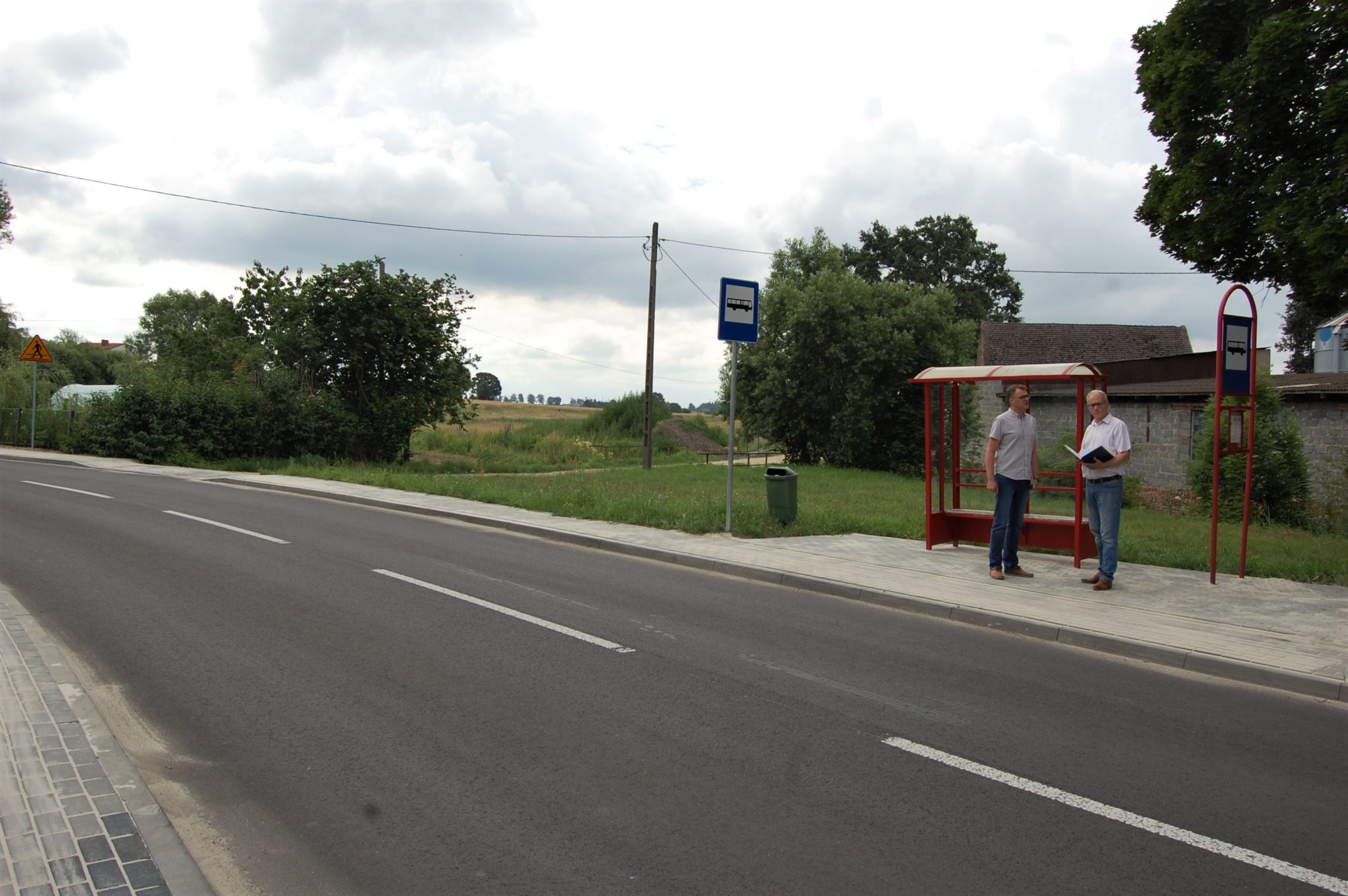 widok na asfaltową drogę z brukowanym chodnikiem, w oddali dwoje mężczyzn przy przystanku autobusowym