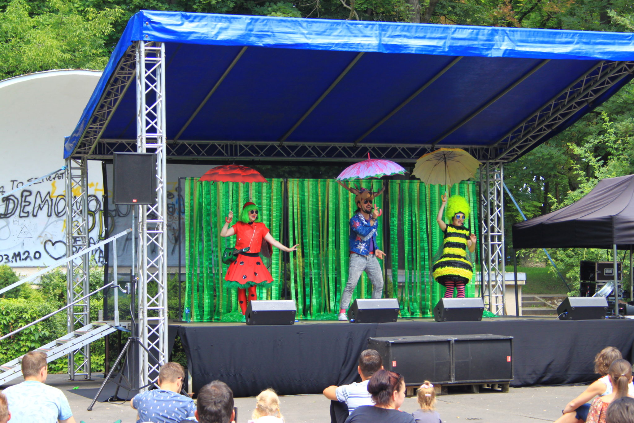 trzy osoby stojące na scenie, każda trzyma w dłoni parasolkę
