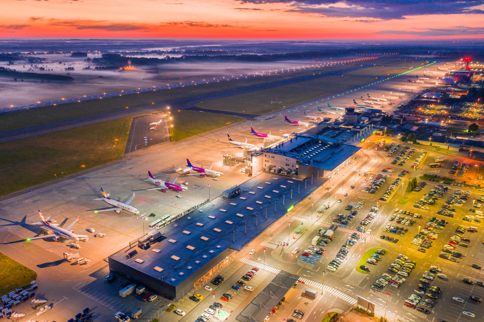 Widok na lotnisko z lotu ptaka. Teren lotniska oświetlony lampami, kilka samolotów stojących na płycie lotniska, w tle kolorowy wschód słońca.