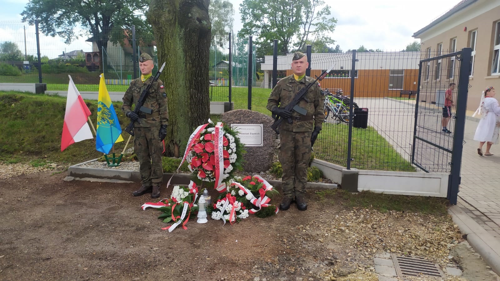 Dwoje mężczyzn w mundurach, którrzy stoją przy pomniku, przed którym leżą wiązanki kwiatów z biało- czerwonymi wstęgami.