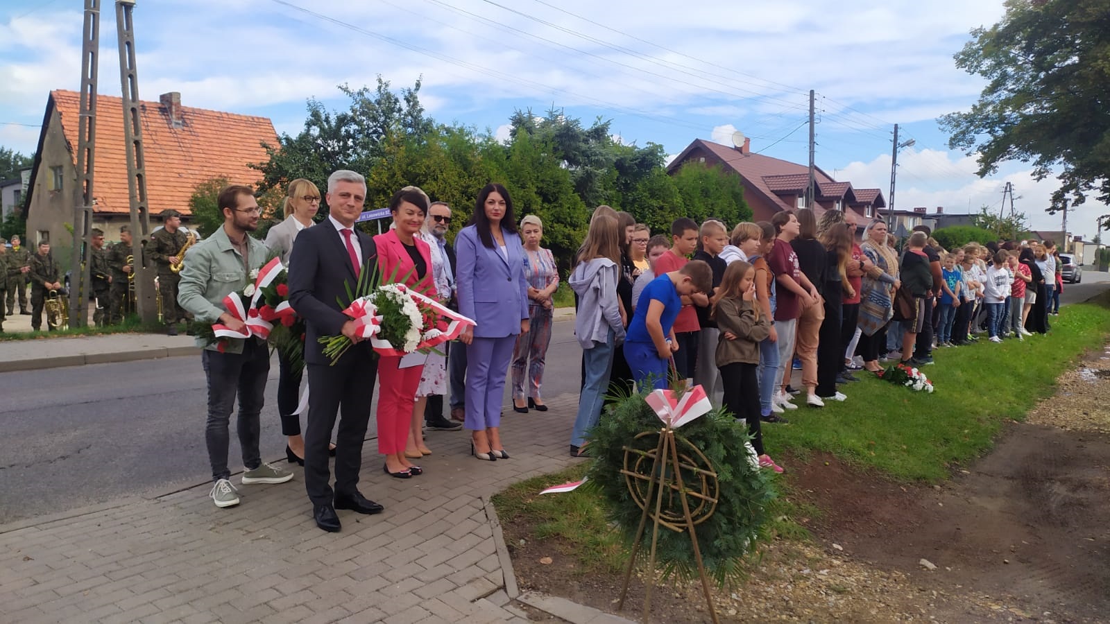 Grupa osób stojących obok siebie. Kilka osób trzyma w rękach wiązanki kwiatów.