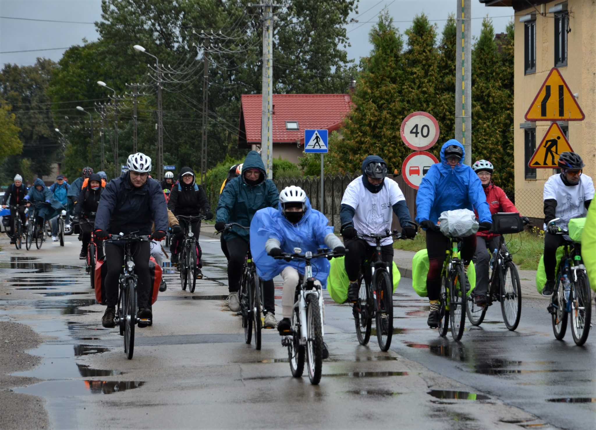 Kilkanaście osób jadących na rowerach, część osób jest ubranych w płaszcze przeciwdeszczowe.
