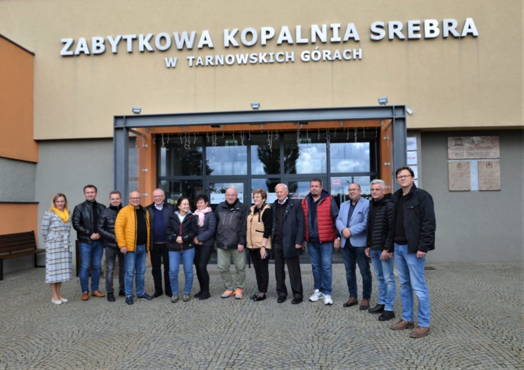 Grupa osób stojących obok siebie, w tle duży budynek z napisem Zabytkowa Kopalnia Srebra w Tarnowskich Górach