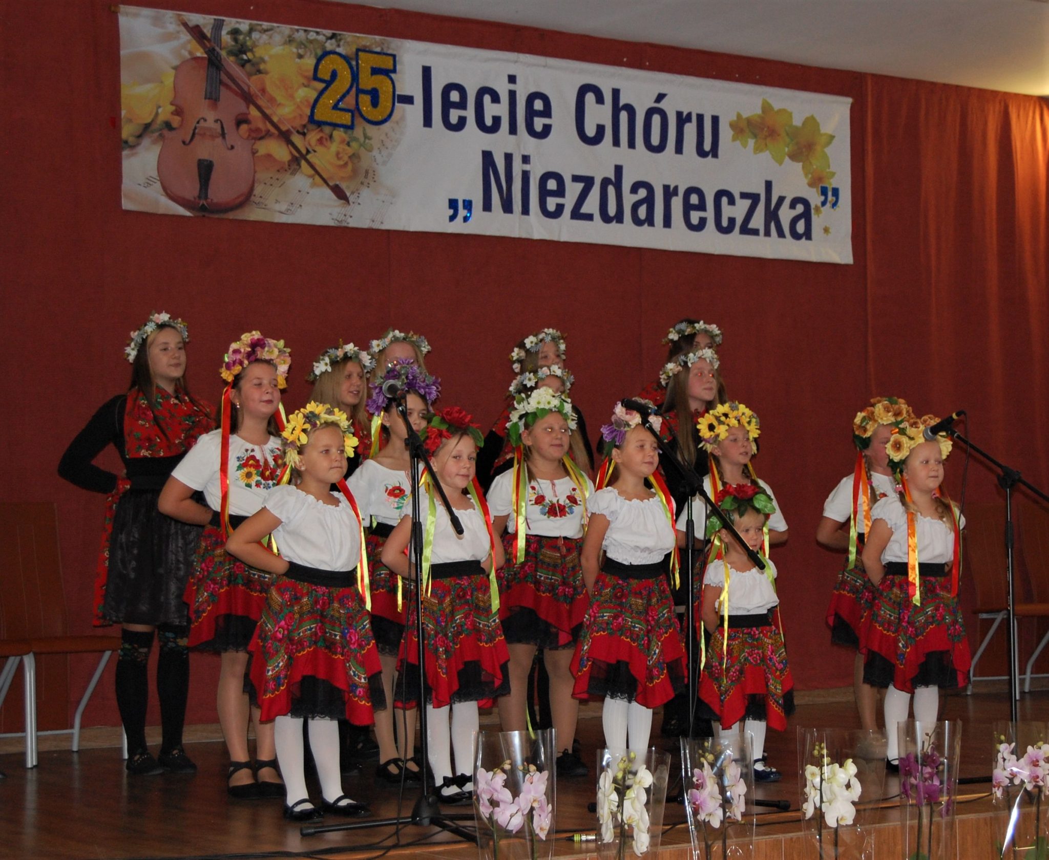 Grupa dziewcząt w strojach regionalnych stojąca na scenie, w tle banek z napisem 25-lecie Chóru Niezdareczka.