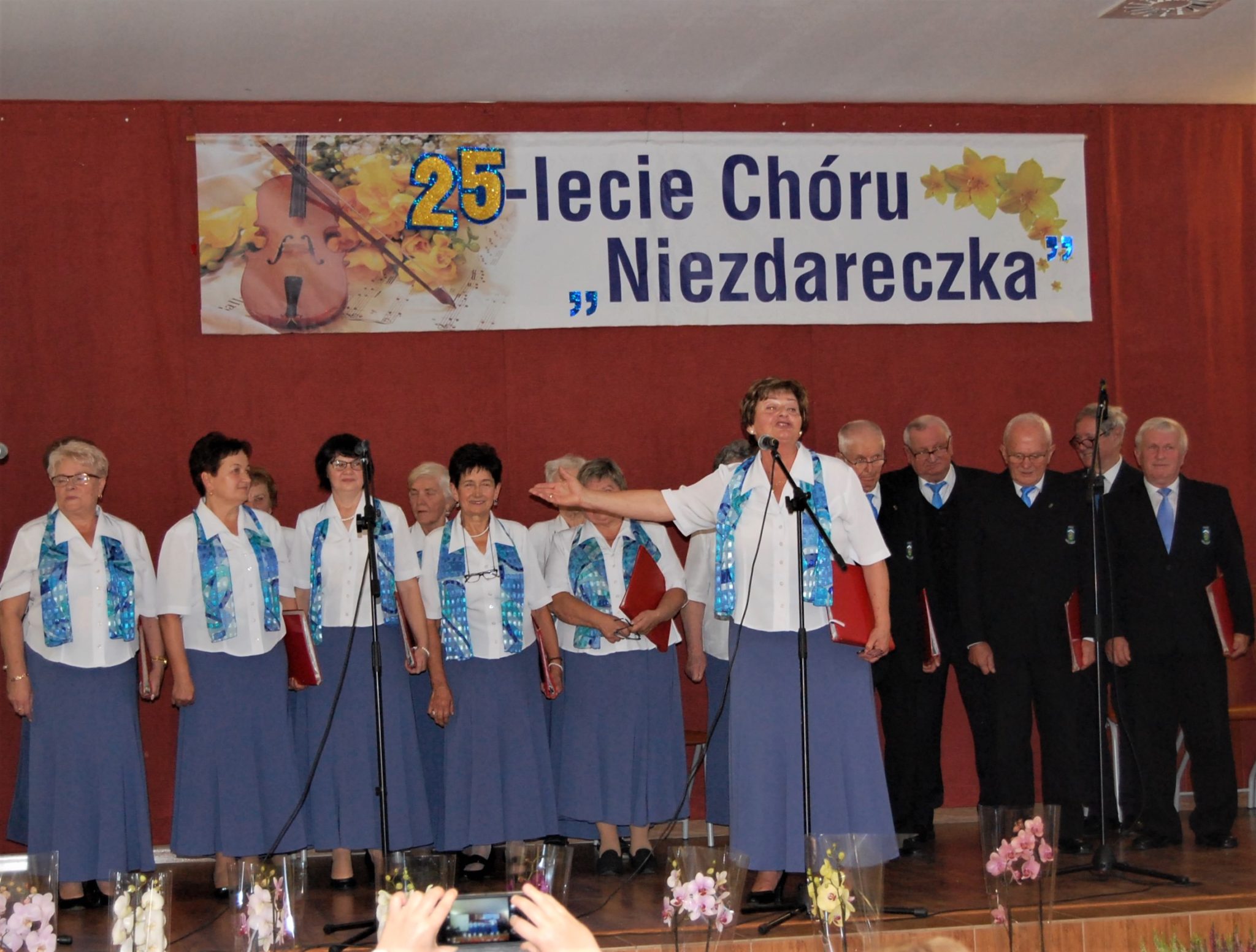 Grupa ludzi stojąca na scenie, mężczyźni ubrani są w garnitury. Kobiety ubrane w niebieskie spódnice i białe bluzki. W tle baner z napisem 25-lecie Chóru Niezdareczka.