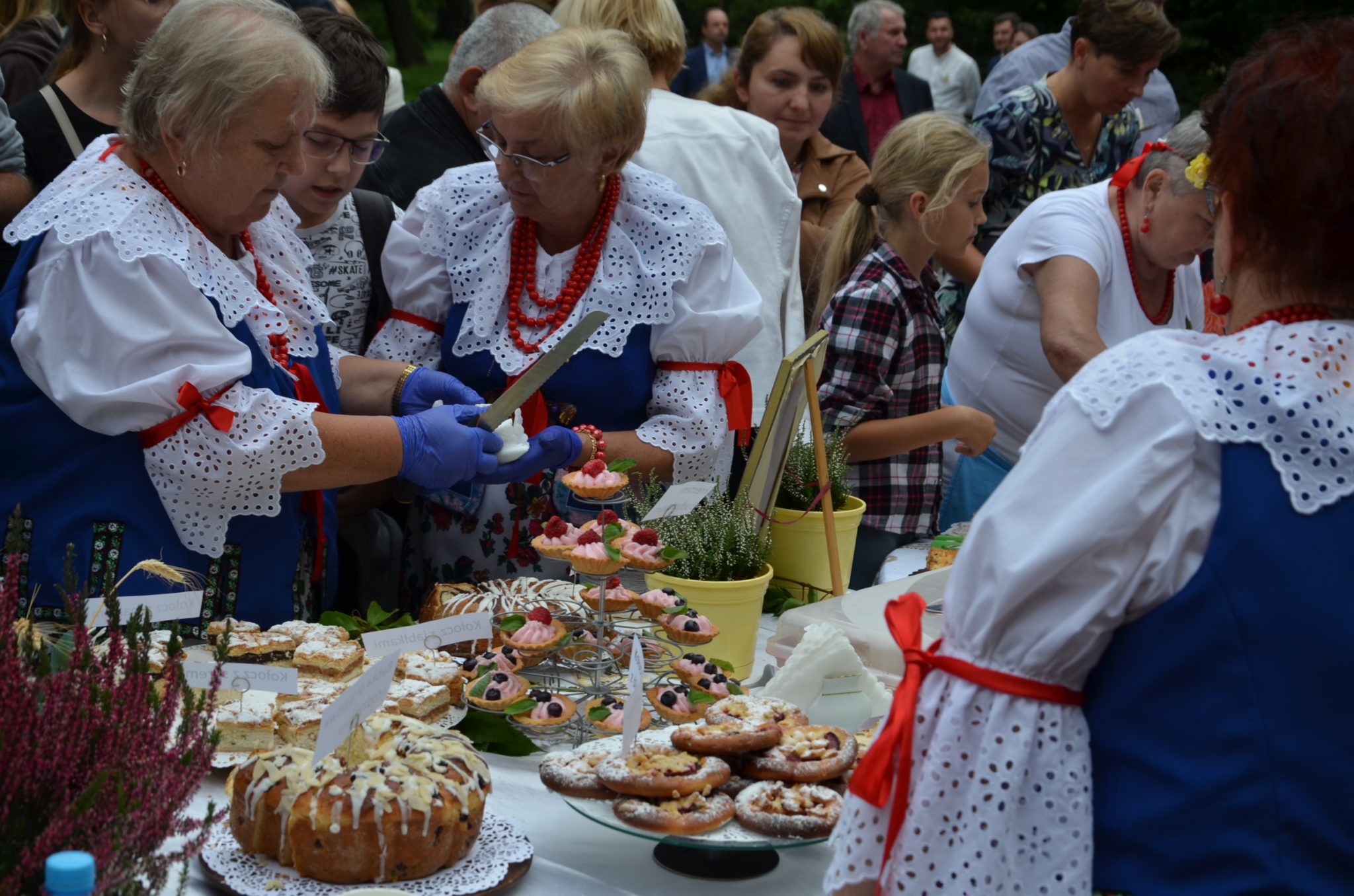 Kilka kobiet ubranych w stroje ludowe, nakładają na talerzyki ciasto, które znajduje się na zastawionym stole