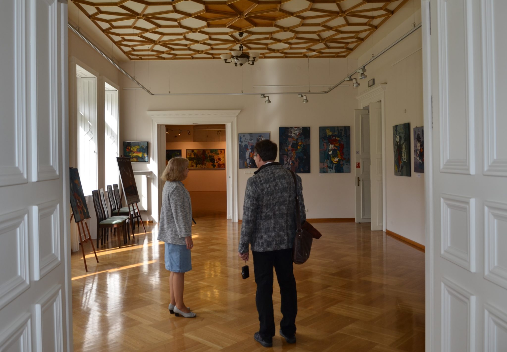 Kobieta i mężczyzna stoją w pomieszczeniu, którego sufit jest dekoracyjnie zdobiony, a na ścianach wiszą obrazy.