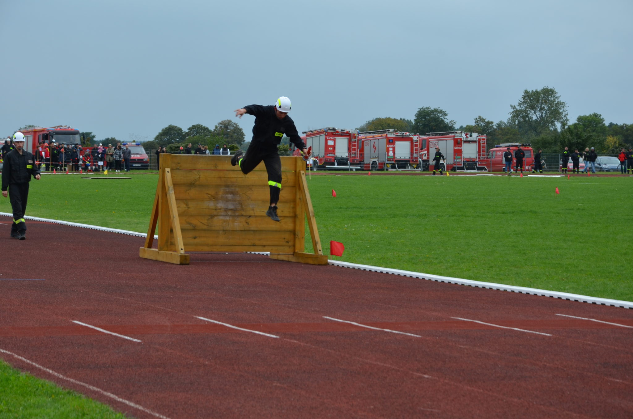 Osoba skacząca przez przeszkodę, w tle boisko sportowe i kilka wozów strażackich.