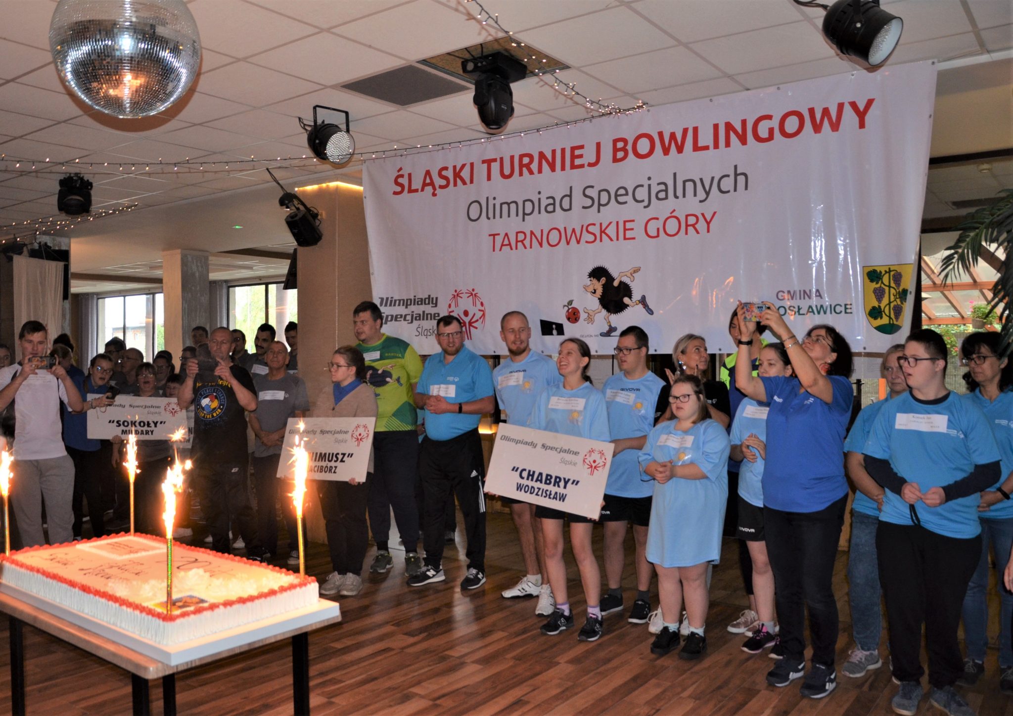 Duża grupa osób stojących obok siebie, za nimi wisi duży banek z napisem Śląski Turniej Bowlingowy Olimpiad Specjalnych Tarnowskie Góry.