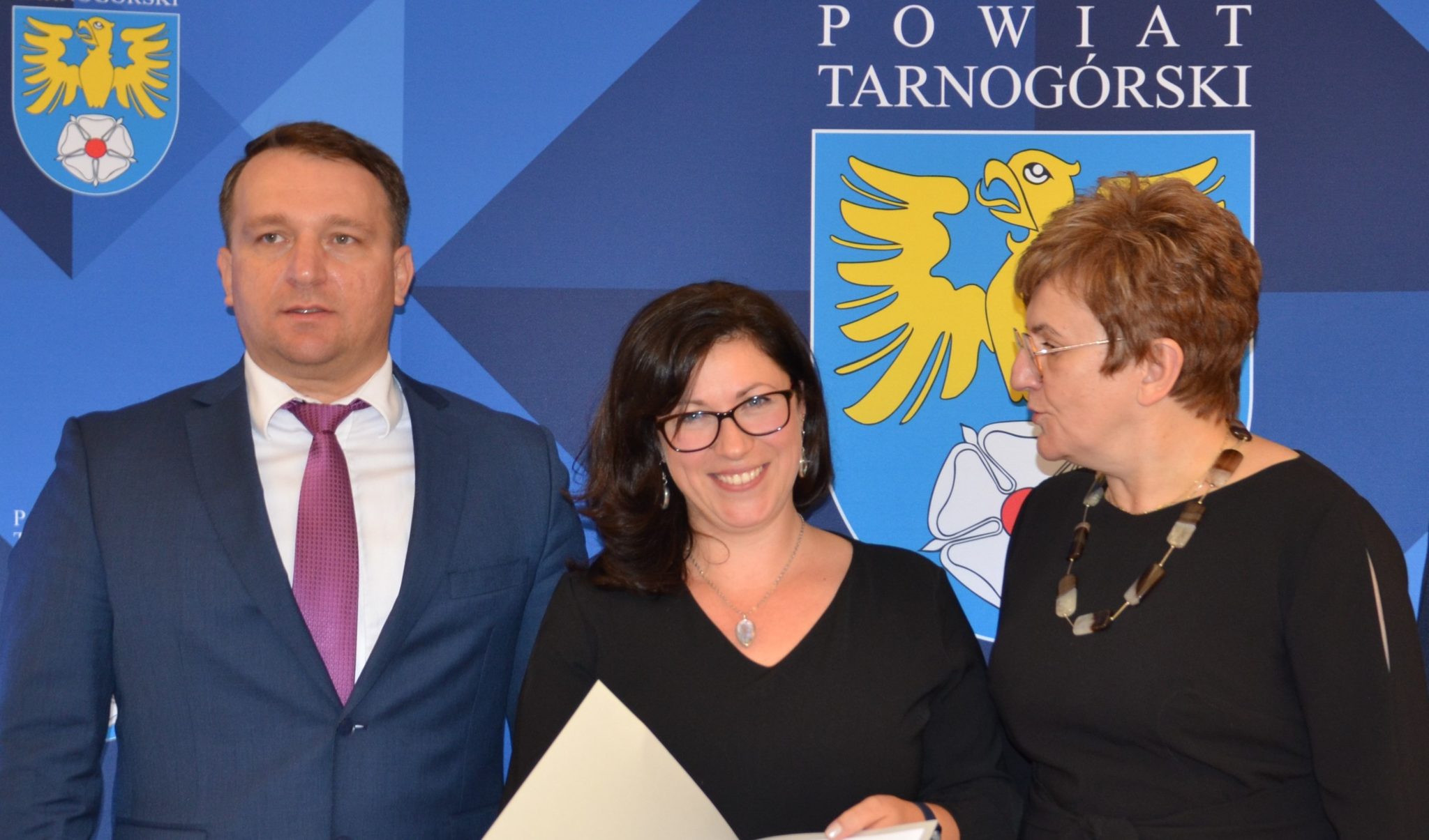 Członkowie Zarządu stojący obok nauczycielki, która pokazuje dokument znajdujący się w ozdobnej teczce.