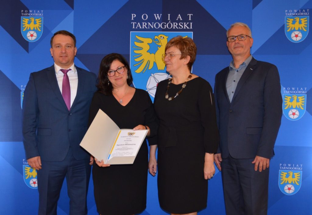 Członkowie Zarządu stojący obok nauczycielki, która pokazuje dokument znajdujący się w ozdobnej teczce.