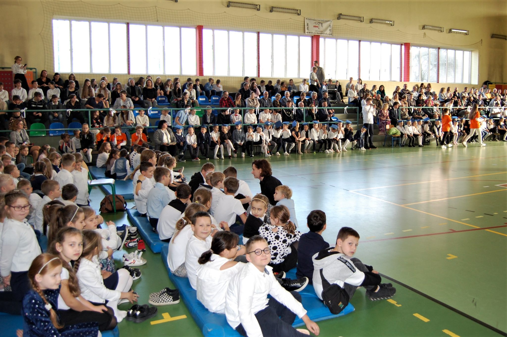 Bardzo duża grupa dzieci siedzących na materacach w sali gimnastycznej, w tle trybuny z pełną widownią.
