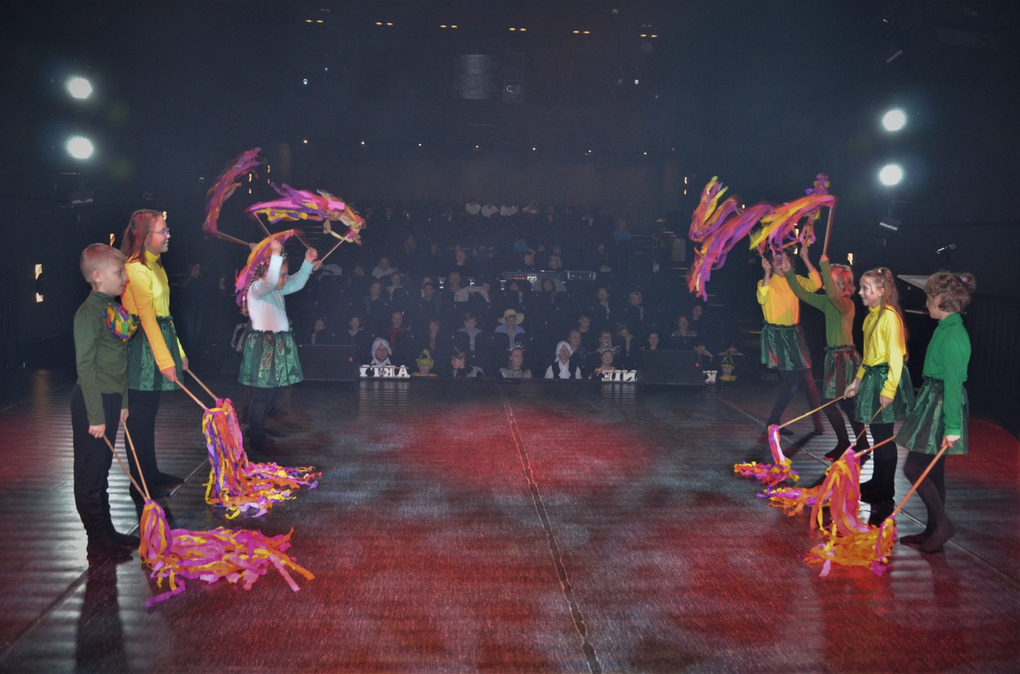 Grupa dzieci na scenie w kolorowych strojach, w tle widoczk na widownę.