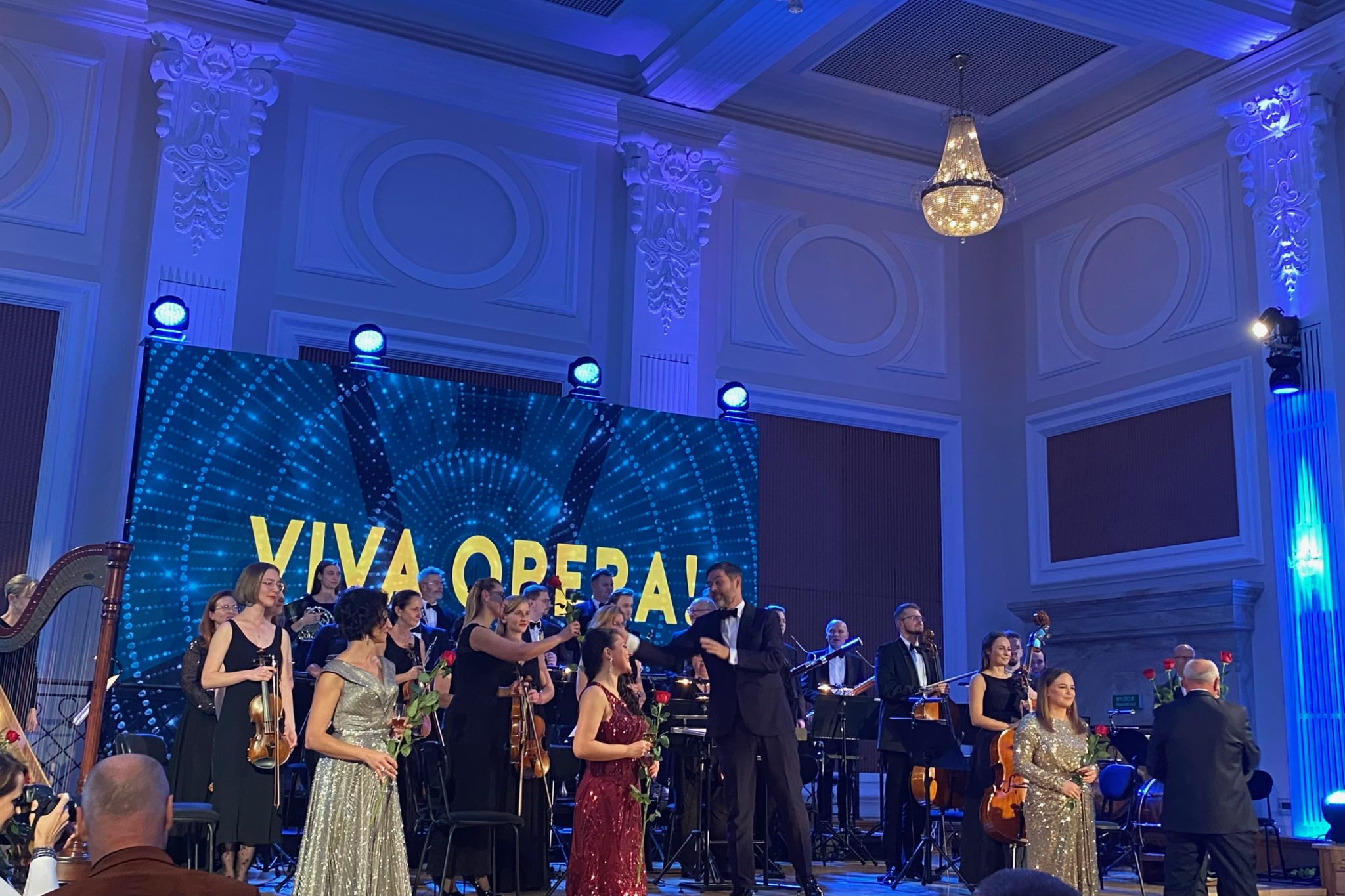 Liczna grupa osób na scenie, na pierwszym planie kobieta w czerwonej sukni. W tle baner z napisem viva opera.