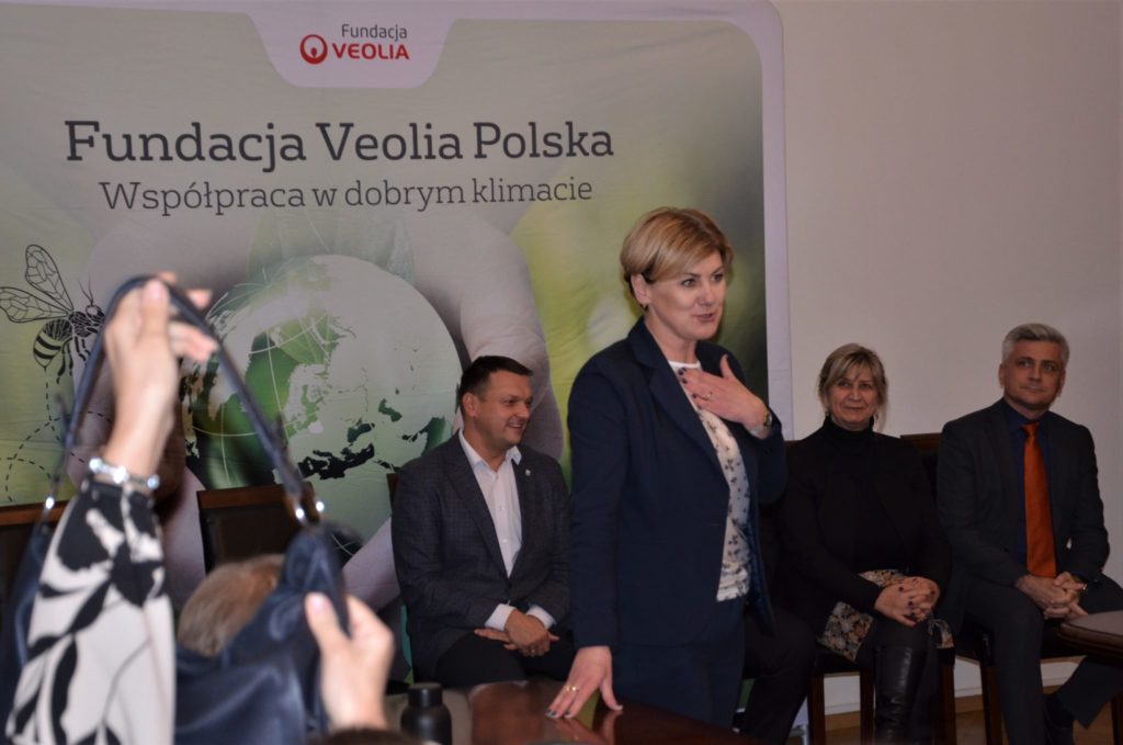Kilka osób znajdujących się w sali, na pierwszym planie kobieta w blond włosach. W tle duży plakat z napisem Fundacja Veolia Polska.