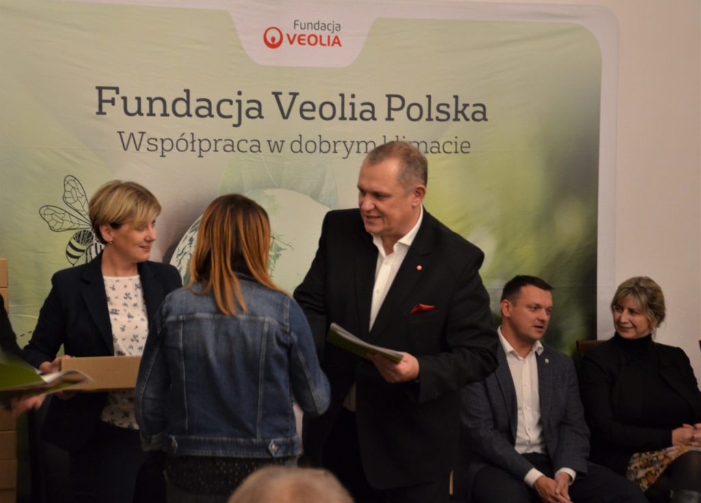 Mężczyzna wręcza dokument kobiecie, w tle plakat z napisem fundacja veolia polska.