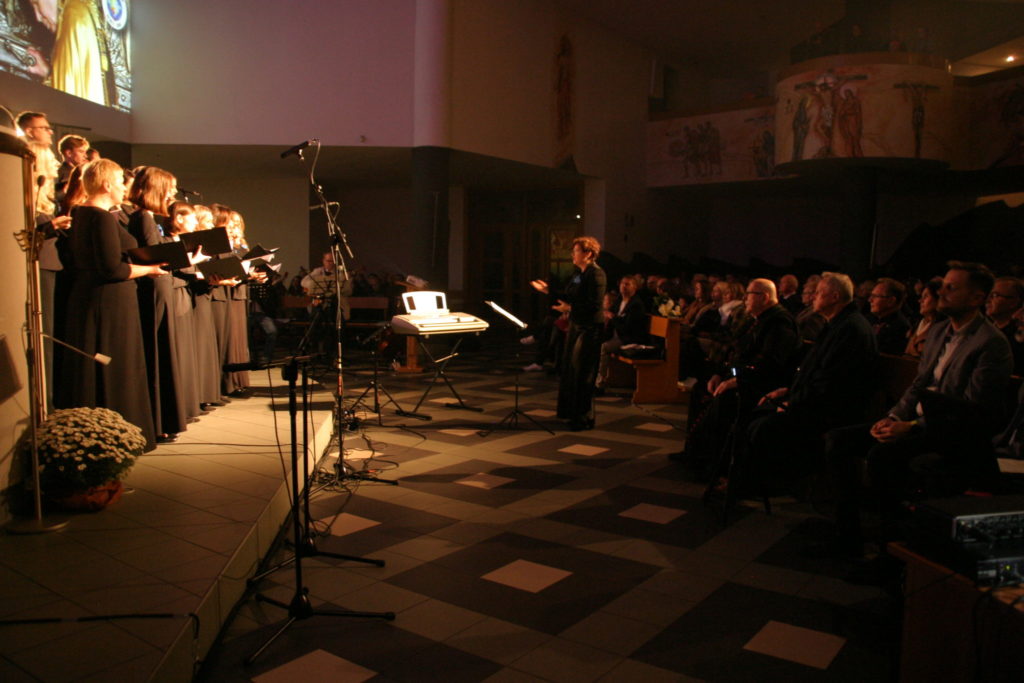 Występ artystyczny w kościele. Grupa osób stoi przed publicznością siedzącą w nawach kościelnych.
