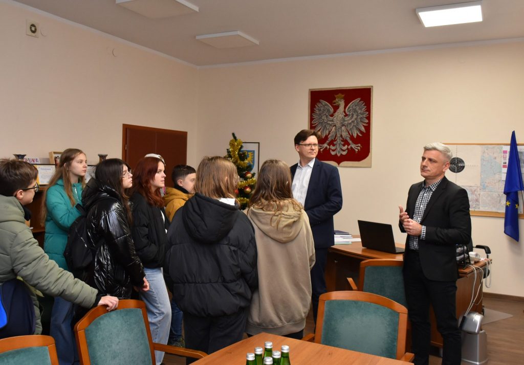 Grupa młodzieży znajduje się w pomieszczeniu, w tle na ścianie wisi Godło Polski.
