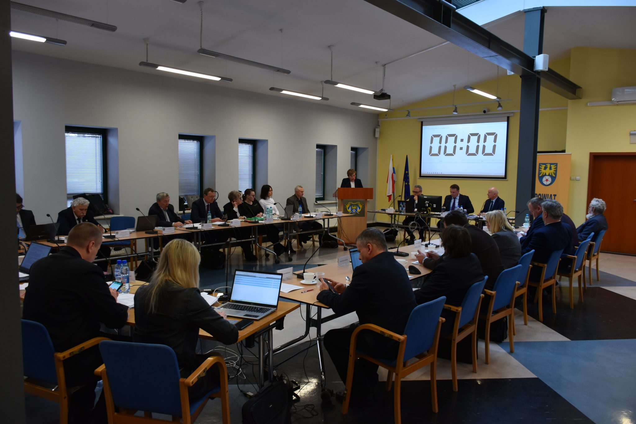 Radni siedzący przy stole podczas sesji. W tle przy mównicy stoi starosta.