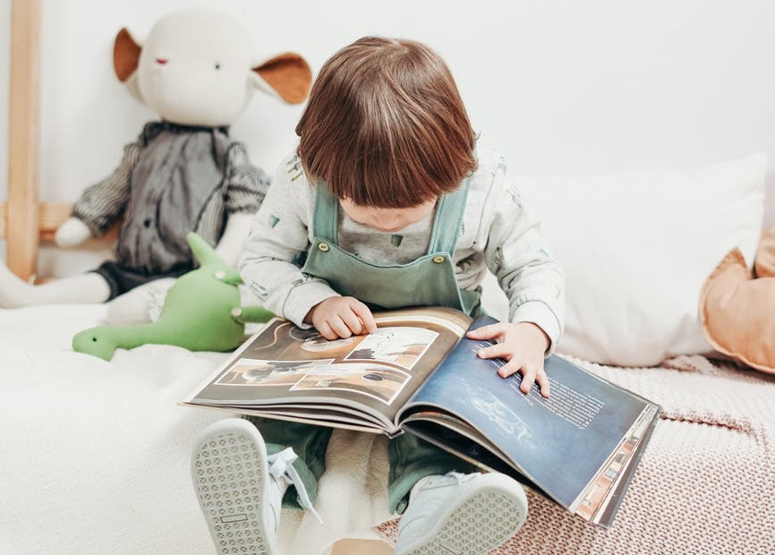 Dziecko, które siedzi na łóżku, na kolanach trzyma książkę. W tle jasna ściana o którą opiera się duża maskotka.