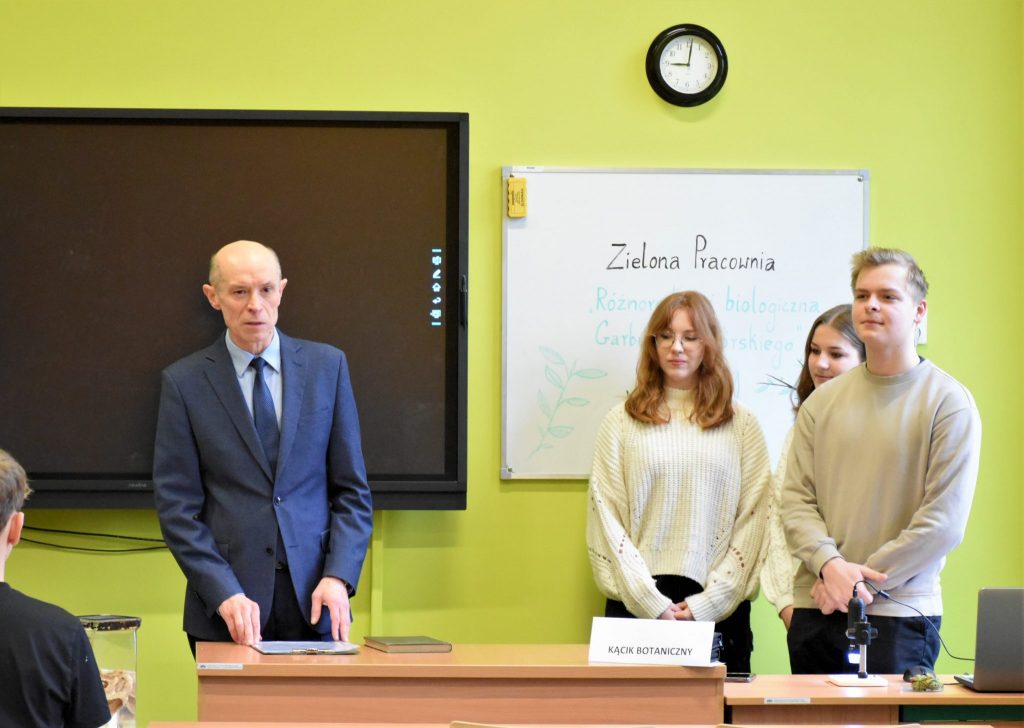 Dyrektor szkoły stojący przy biurku, w tle tablica z napisem Zielona Pracownia.