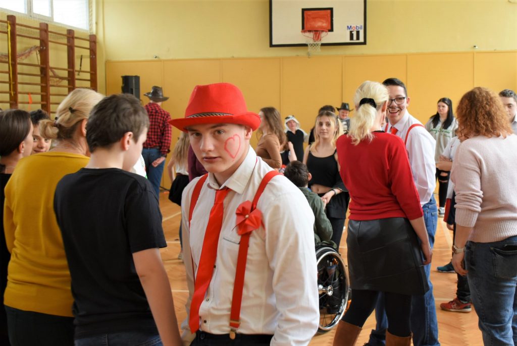 Chłopiec w czerwonym kapeluszu. W tle grupa młodzieży na sali gimnastycznej.