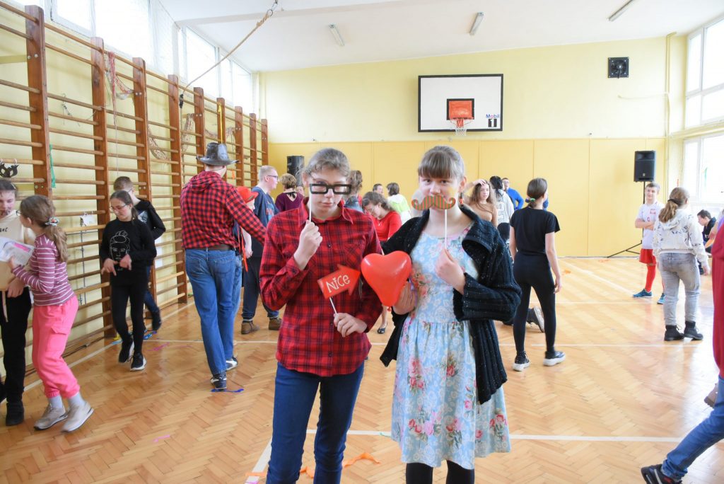 Dzieci podczas zabawy na sali gimnastycznej. Na pierwszym planie dziewczyna i chłopak z akcesoriami do zdjęć: okularami, wąsem i balonem.