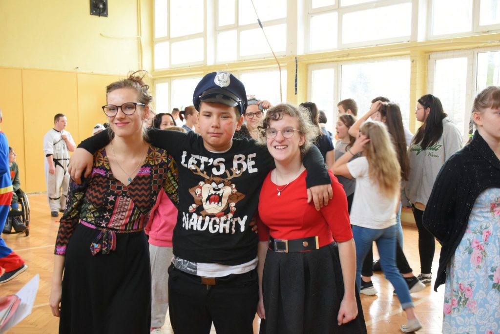 Troje dzieci stojących obok siebie. Dwie dziewczynki, a między nimi chłopiec w kapeluszu. W tle dzieci rozmieszone w dużym pomieszczeniu.