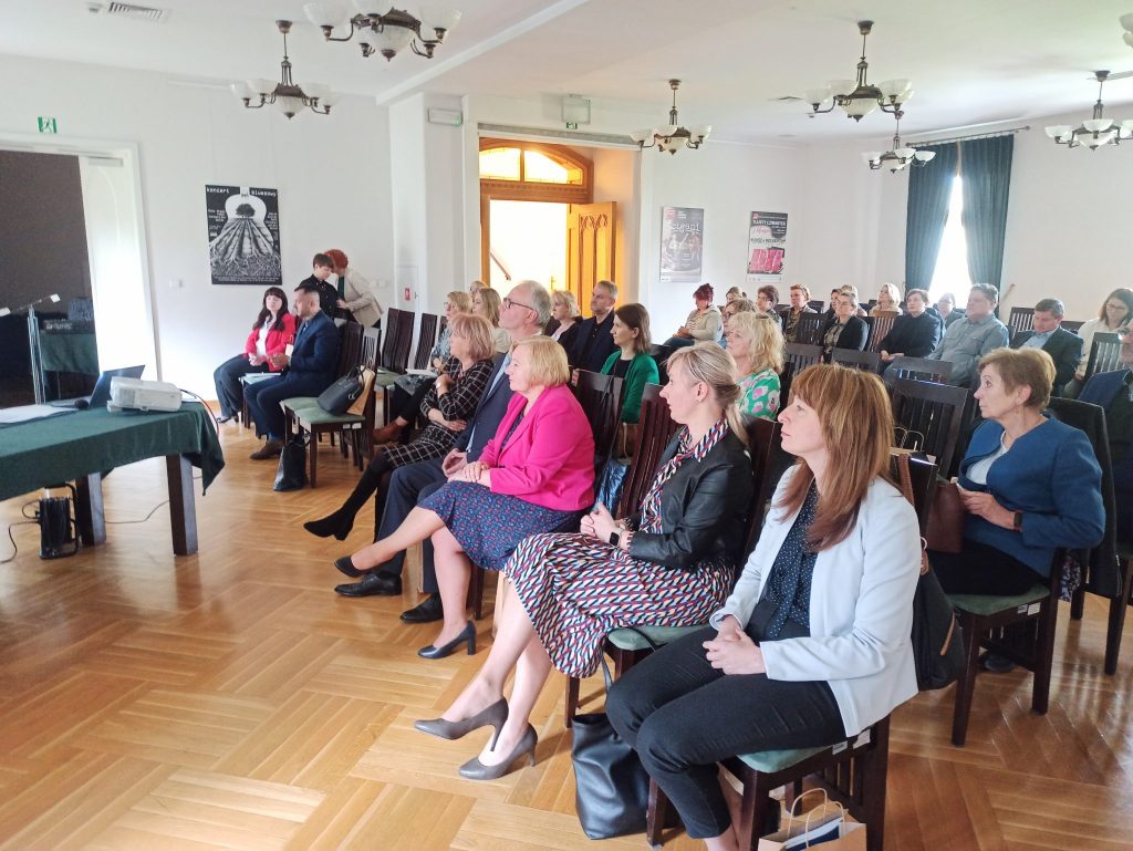 Grupa osób zgromadzona w dużej sali. w tle ściana z dużym oknem oraz duże drzwi skrzydłowe.