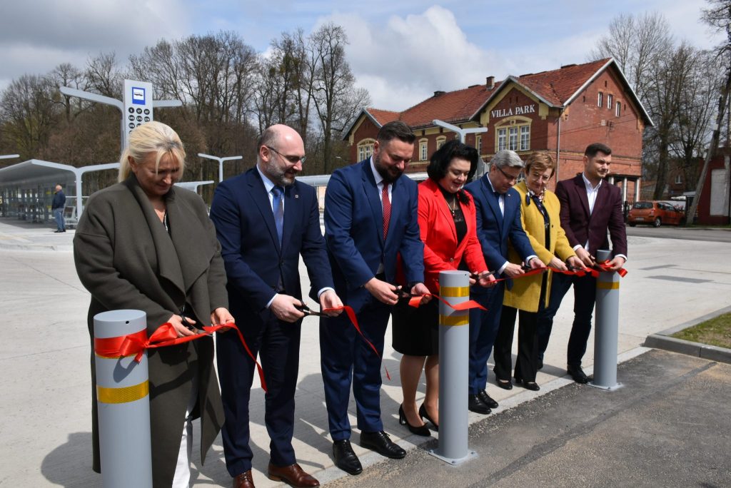 Kilka osób stojących obok siebie podczas przecinania czerwonej wstęgi. W tle centrum przesiadkowe.