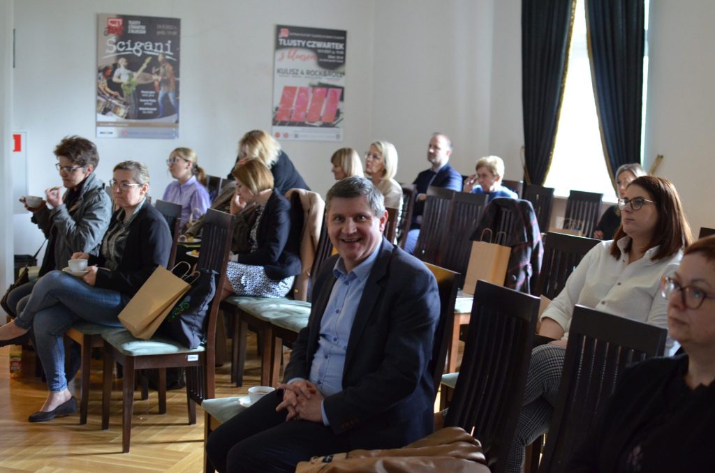 Grupa osób siedząca na stołkach w dużym pomieszczeniu. Na pierwszym planie uśmiechnięty mężczyzna.