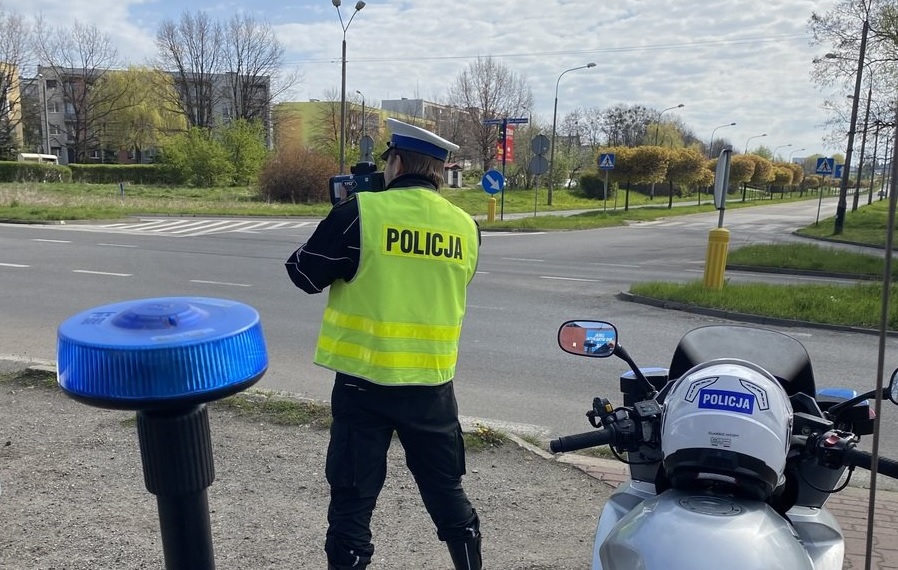 Osoba w kamizelce z napisem policja, obok stoi motocykl. W tle widoczna droga i zabudowa miejska.