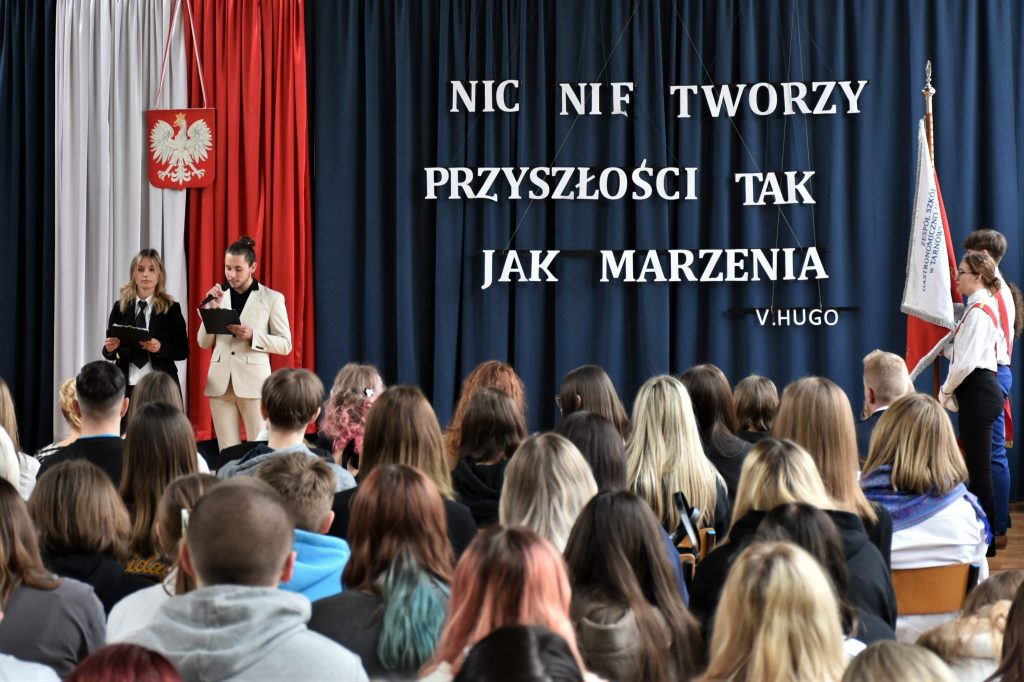 Grupa osób zgromadzona w sali podczas akademii. W tle napis: nic nie tworzy przyszłości tak jak marzenia.