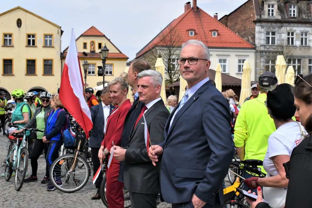 Przedstawiciele zarządu powiatu, którzy trzymają w dłoniach flagi polski. W tle zgromadzeni mieszkańcy oraz rowerzyści.