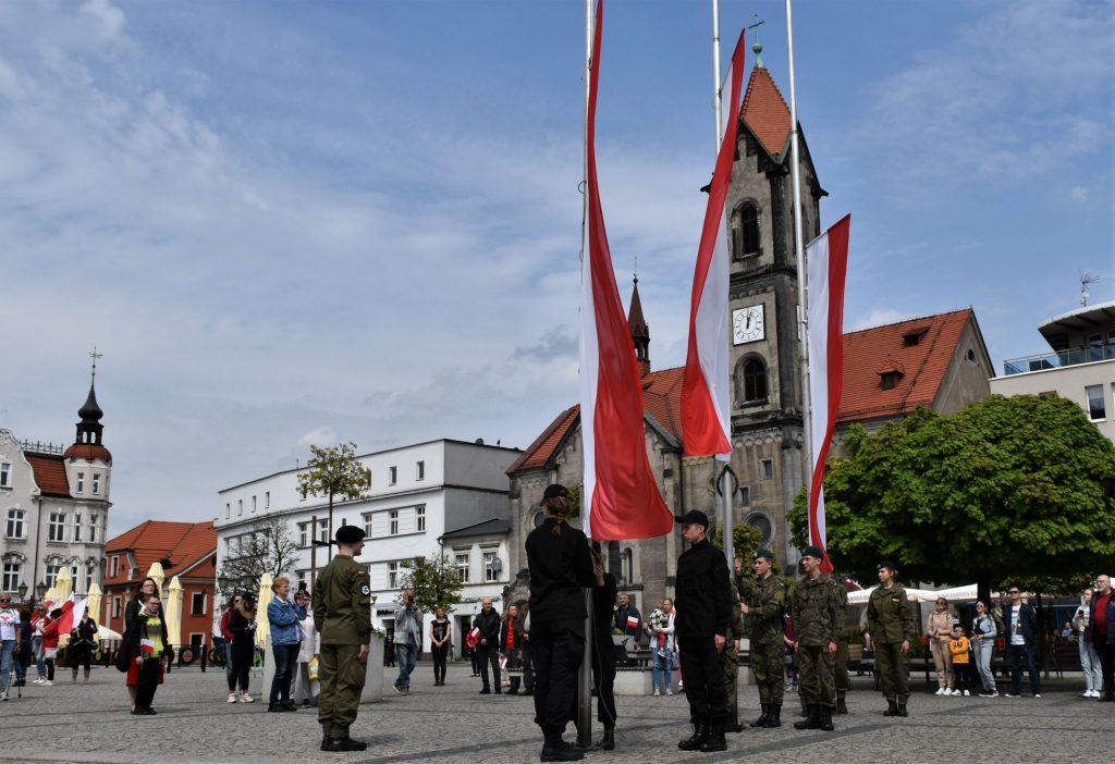 Flagi wciągane na maszt. W tle rozproszona duża grupa ludzi.