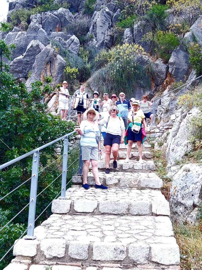 Grupa osób stojących na schodach, w tle duży masyw skalny.