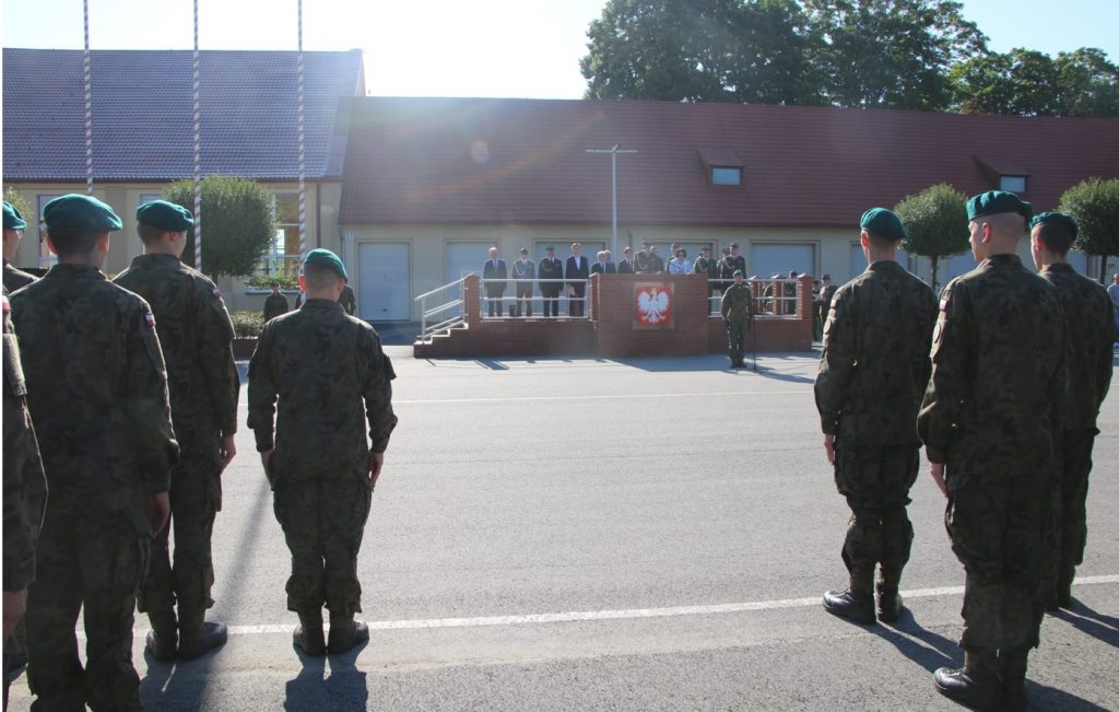 Grupa osób zgromadzona na placu, na pierwszym planie żołnierze w mundurach.